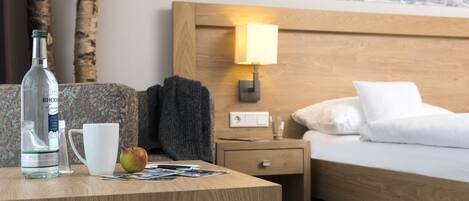 A hotel room with a wooden headboard, a bedside lamp, a bed with white bedding, a nightstand with a lamp, a wooden table with a bottle, a mug, and an apple.