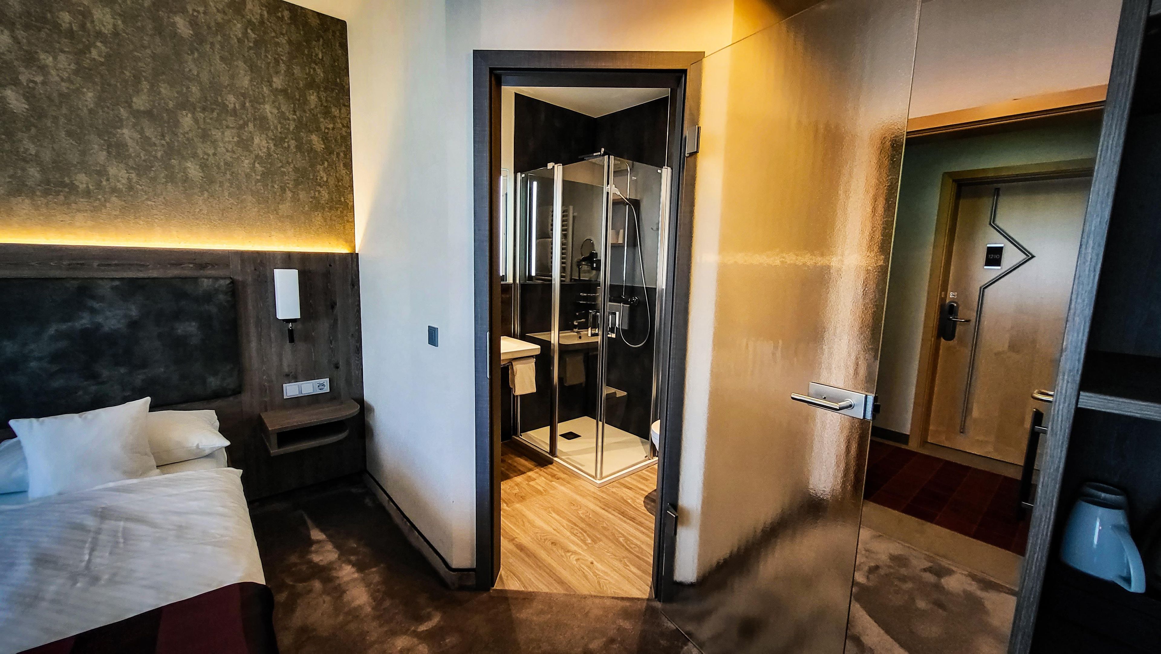 A hotel room with a bed, a bedside table, a nightstand, a mirror, a wall-mounted light, and a door leading to a bathroom with a shower and a toilet.