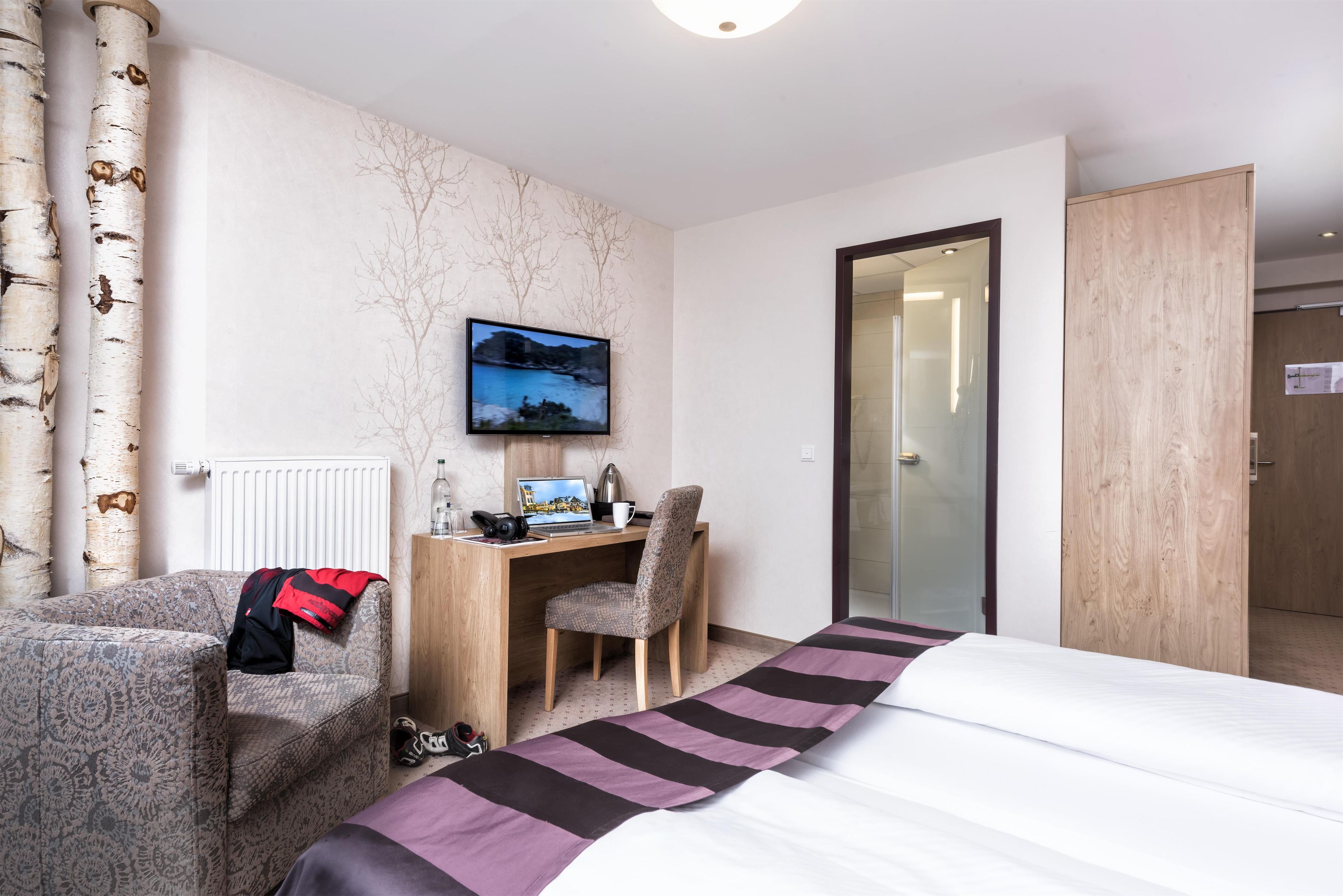 Ein Hotelzimmer mit einem Bett, einem Schreibtisch mit Computer, einem Stuhl, einem an der Wand befestigten Fernseher und einem Fenster.