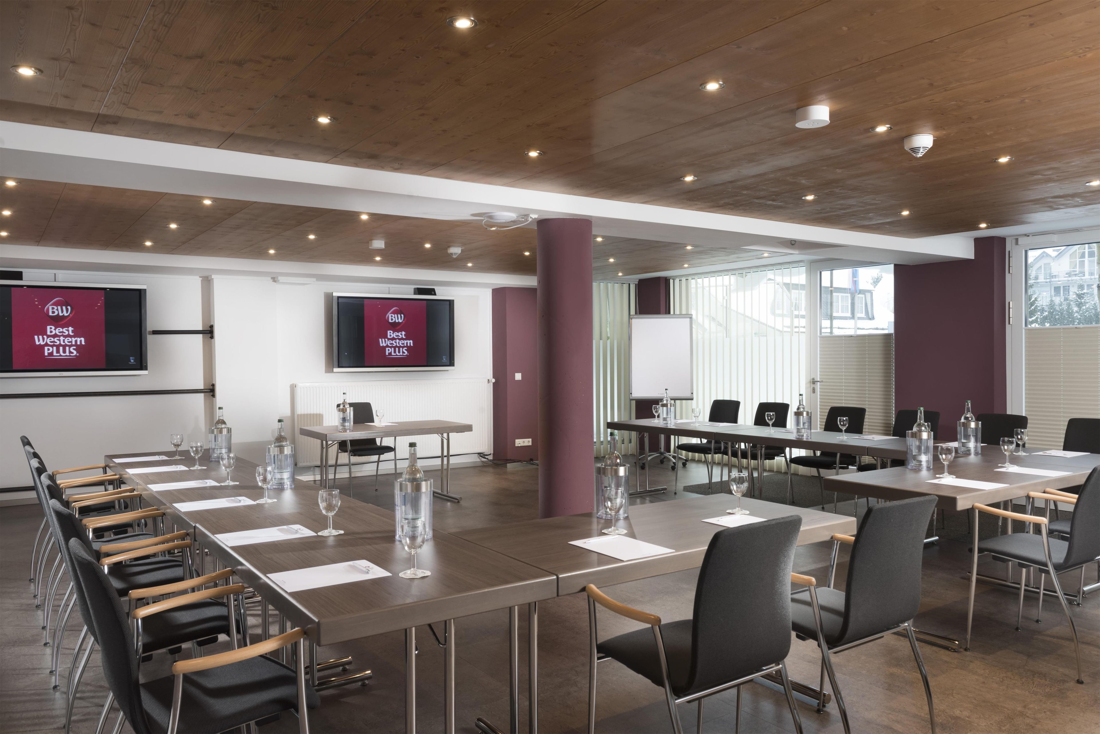 Un salón de conferencias con varias mesas y sillas, equipado con botellas de agua y vasos, y una pantalla grande que muestra el logotipo de 'Best Western PLUS'.