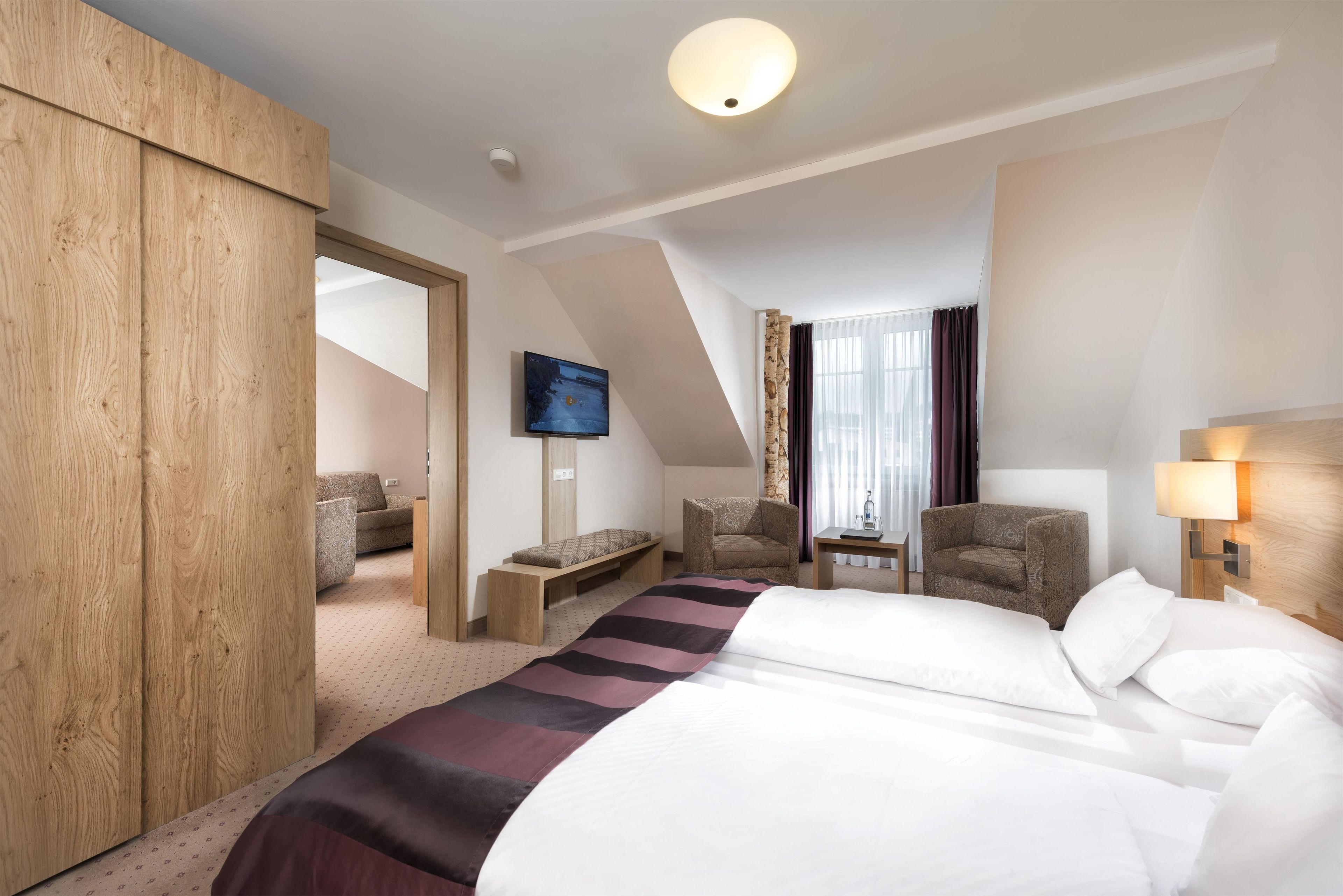 Habitación de hotel con cama, una zona de estar con un banco, una silla, un televisor montado en la pared y una ventana con cortinas.