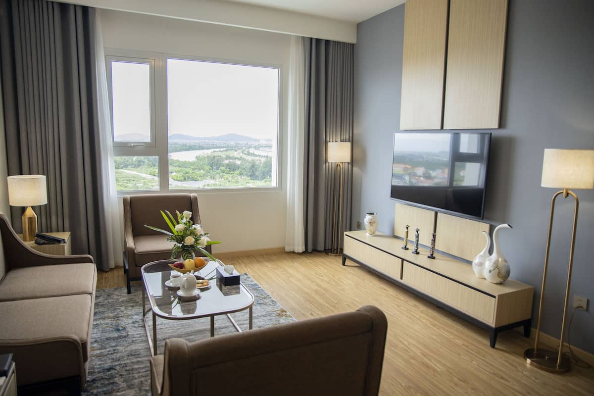 A modern hotel room with a large window offering a scenic view, a flat-screen TV, a sofa, a coffee table with decorative items, and a sideboard with lamps.