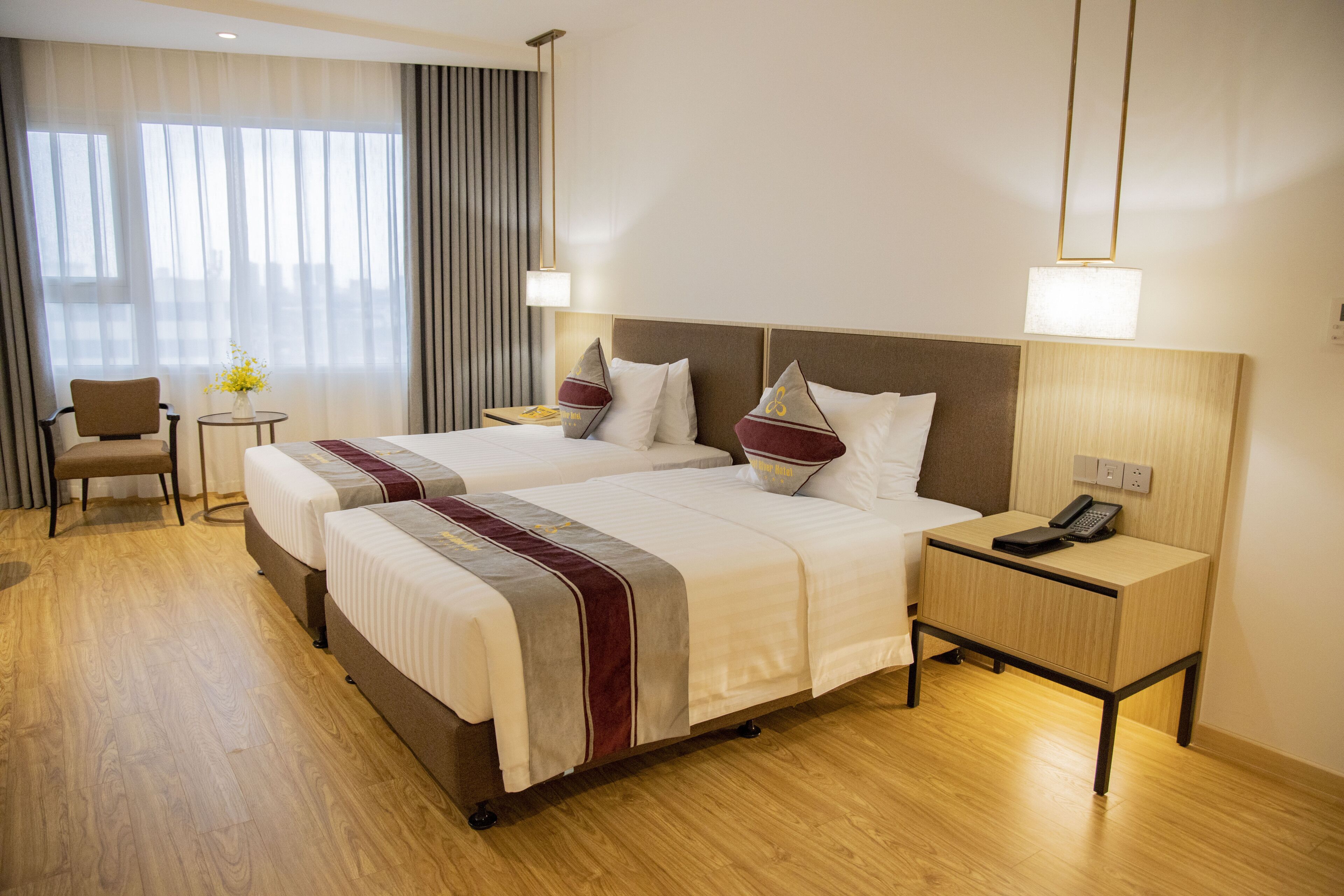 A hotel room with two beds, a small table with a vase of flowers, a chair, and a bedside table with a phone.