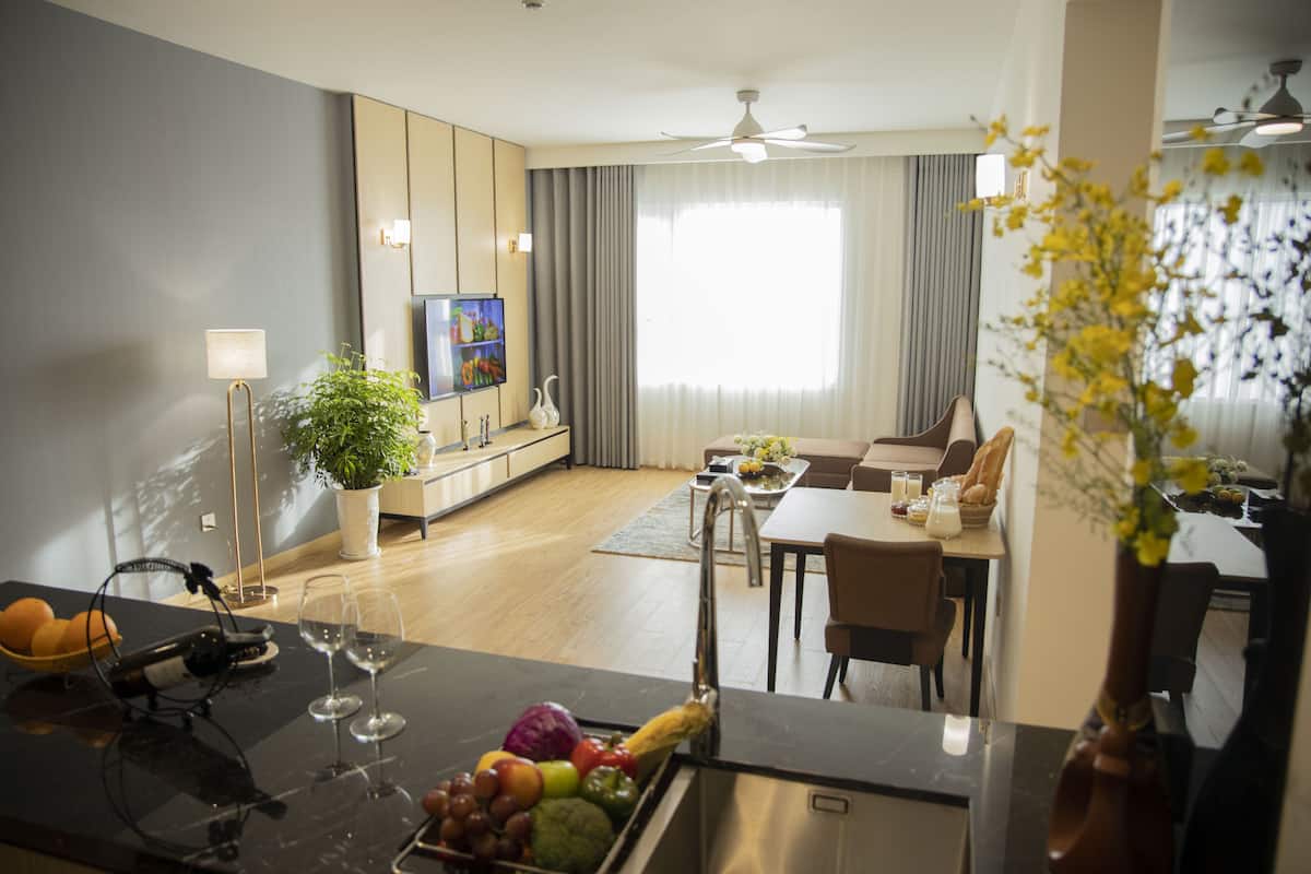 A modern living room with a dining area, a kitchen with a fruit bowl, and a large window with curtains.