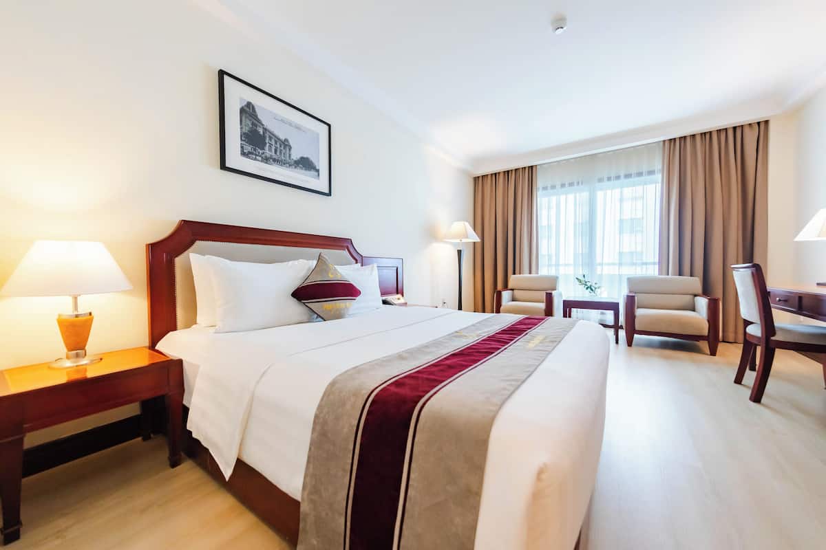 A hotel room with a large bed, two bedside tables with lamps, a desk, two chairs, a framed picture on the wall, and a window with curtains.