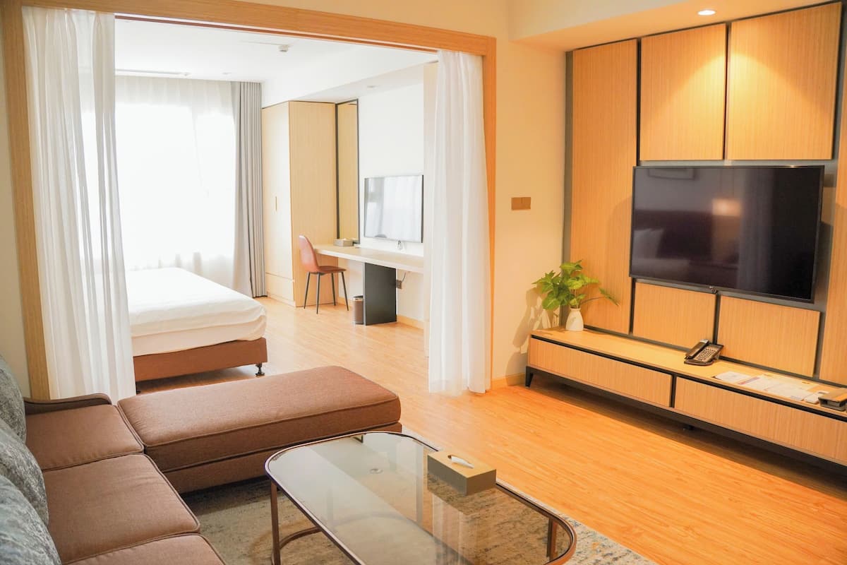 A modern hotel room with a sofa, a glass coffee table, a TV, a desk, and a bed.