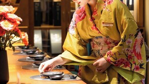 A person in traditional attire arranging dishes on a table in a warmly lit room with wooden paneling and shelves in the background. - Pearl River Hotel (Hai Phong)