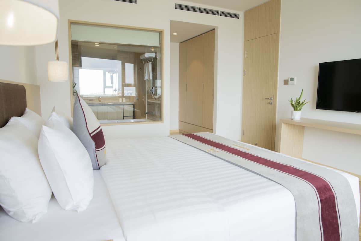 A hotel room with a large bed, a TV mounted on the wall, and a view of the bathroom through a glass door.