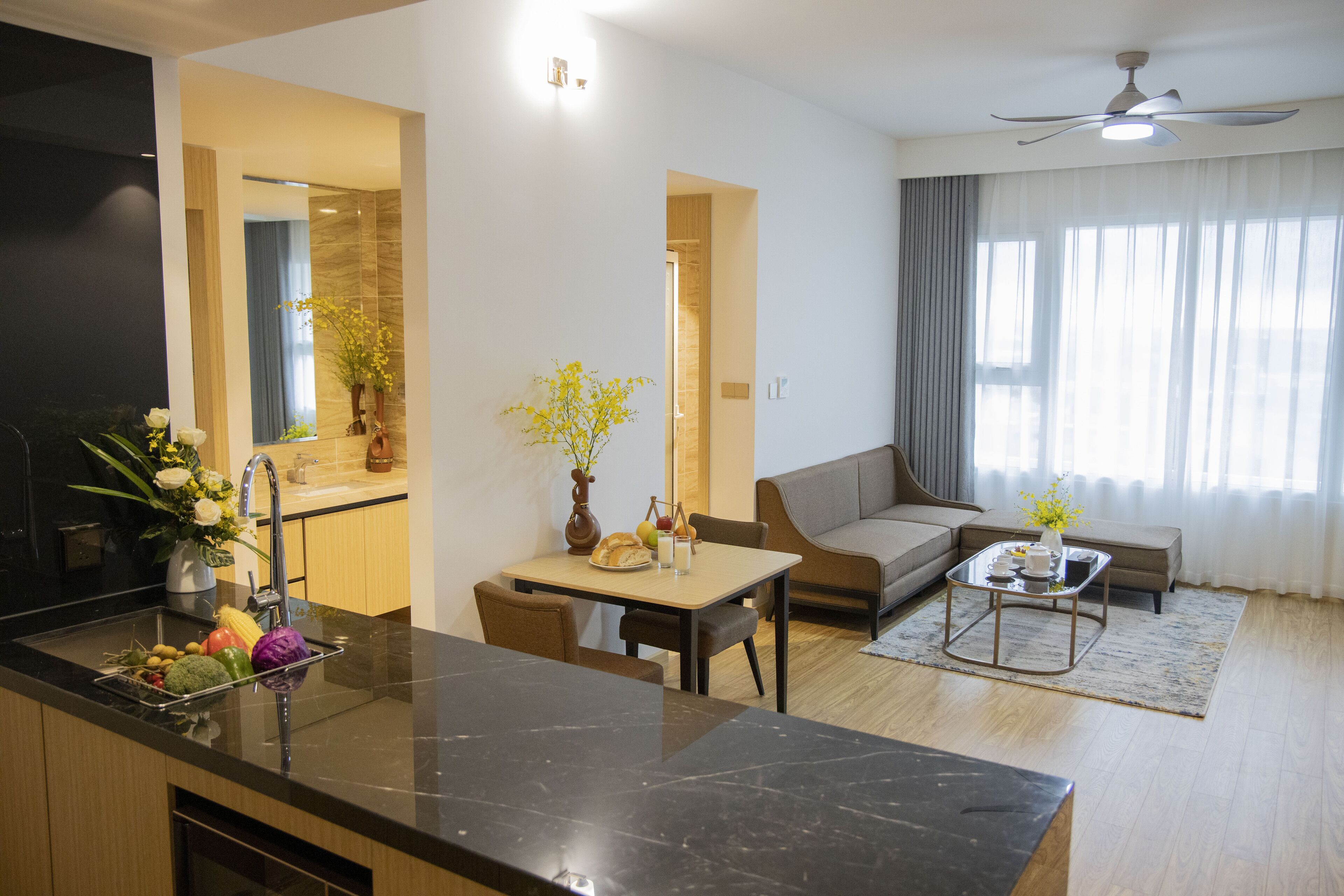 Eine moderne Küche mit Marmorarbeitsplatte, ein Essbereich mit Tisch und Stühlen, ein Sofa und ein Couchtisch. Ein Deckenventilator und ein Fenster mit Vorhängen sind ebenfalls vorhanden.