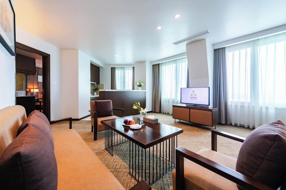 A modern hotel room with a living area, a television, a dining table with a vase of flowers, and a sofa.