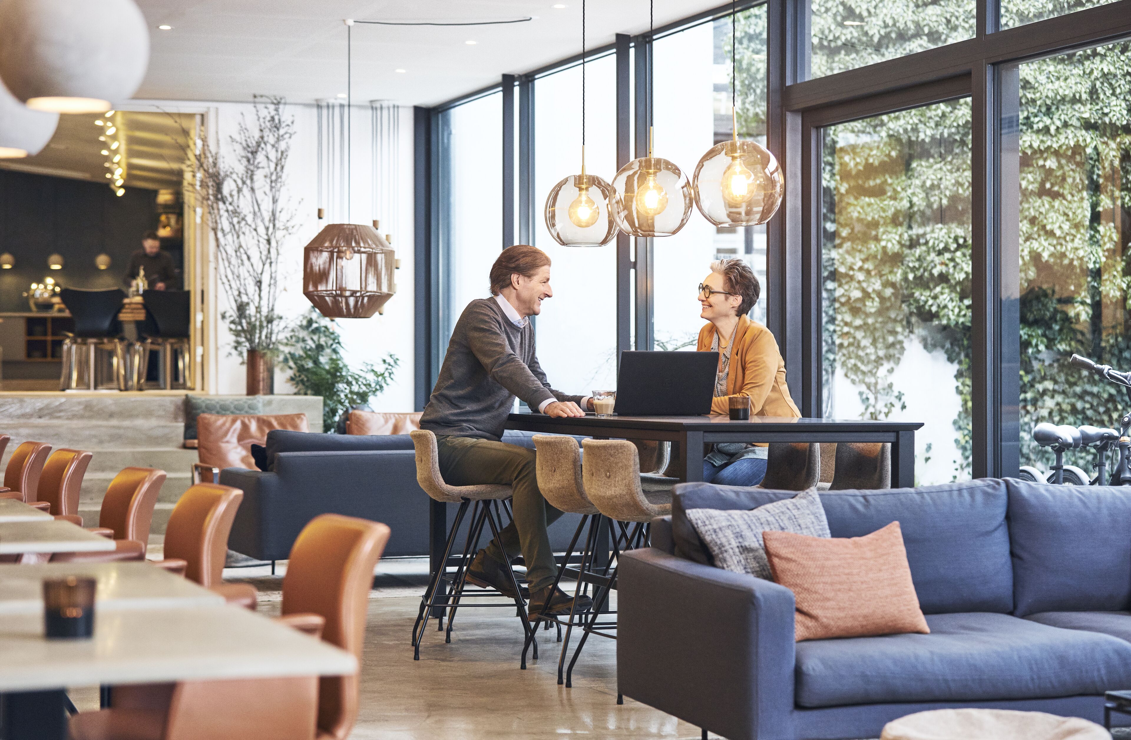 A modern cafe with a large window, a blue sofa, and a table with two people working on laptops.