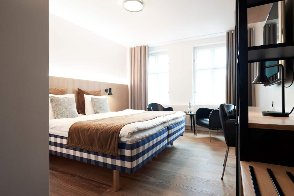 Une chambre d’hôtel avec un grand lit, un bureau avec une lampe, une chaise et une fenêtre avec des rideaux.