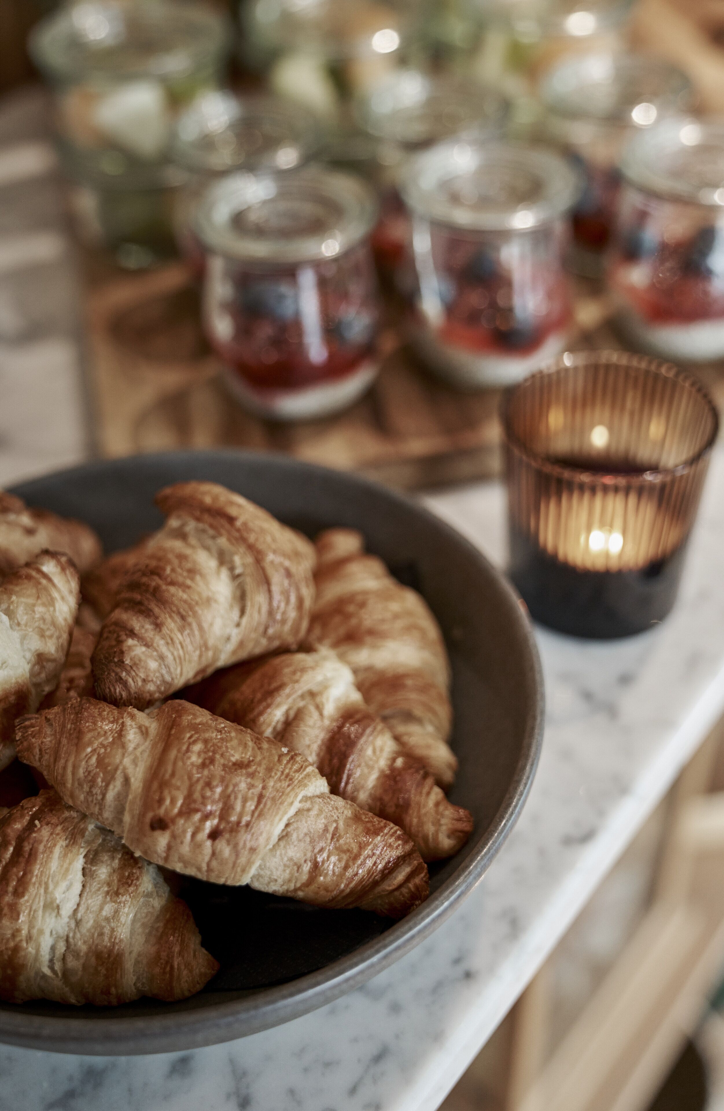 A bowl of croissants on a marble countertop with jars of jam and a candle in the background.