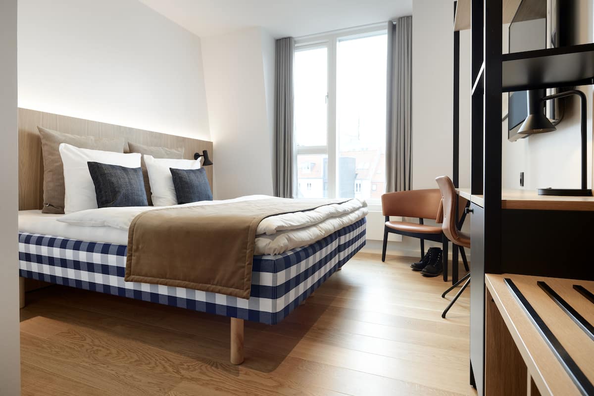 Une chambre d’hôtel moderne avec un lit à motifs à carreaux, un bureau en bois et une vue sur la ville grâce à de grandes fenêtres.