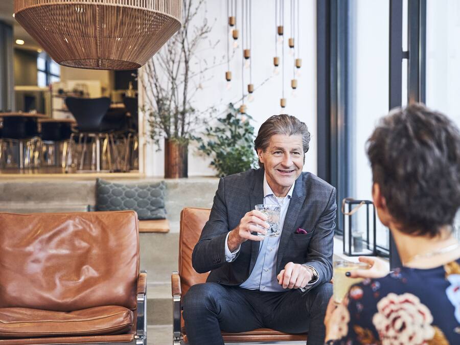 Un hombre con traje sentado en un sillón de cuero, sosteniendo una copa, en un ambiente interior moderno con un ventanal grande y lámparas colgantes.