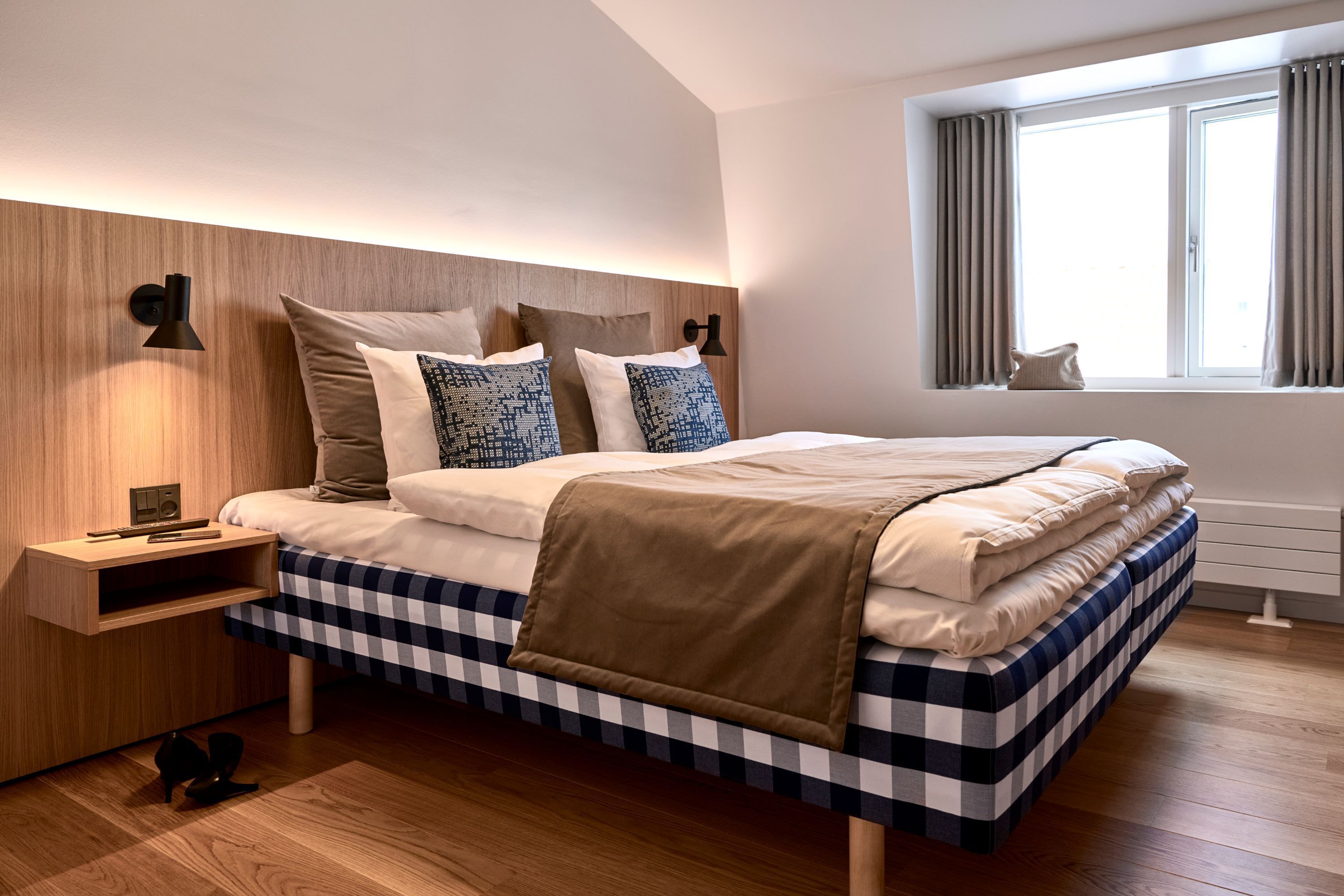 A modern bedroom with a checkered bedspread, a wooden headboard, bedside lamps, and a window with curtains.