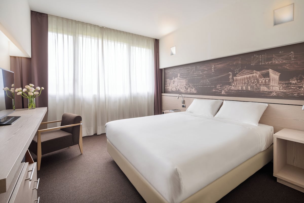 Ein Hotelzimmer mit einem großen Bett, einem Schreibtisch, einem Sessel, einem Nachttisch und einem Wandbild.