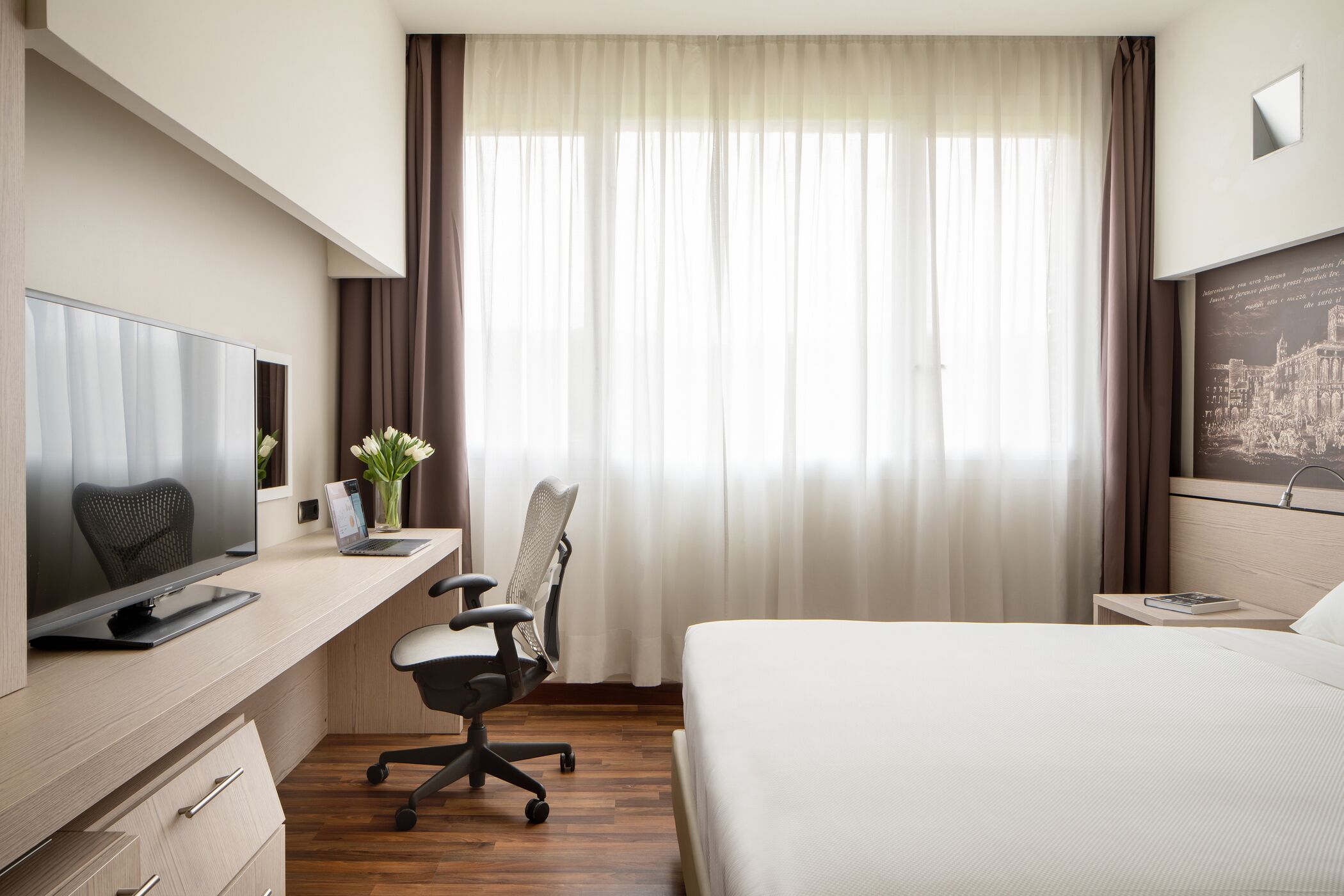 Une chambre d’hôtel avec un lit, un bureau équipé d’un ordinateur, une chaise, une télévision et une fenêtre avec des rideaux.