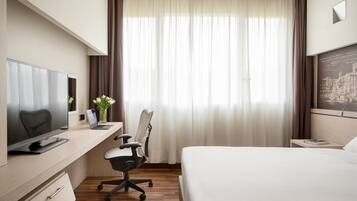 Une chambre d’hôtel avec un lit, un bureau équipé d’un ordinateur, une chaise, une télévision et une fenêtre avec des rideaux.