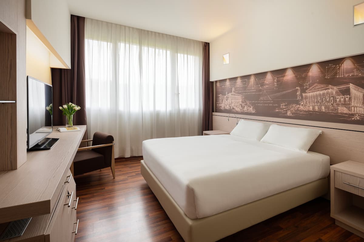 Ein modernes Hotelzimmer mit einem großen Bett, einem Schreibtisch mit Computer, einem Sessel und einem an der Wand befestigten Fernseher.