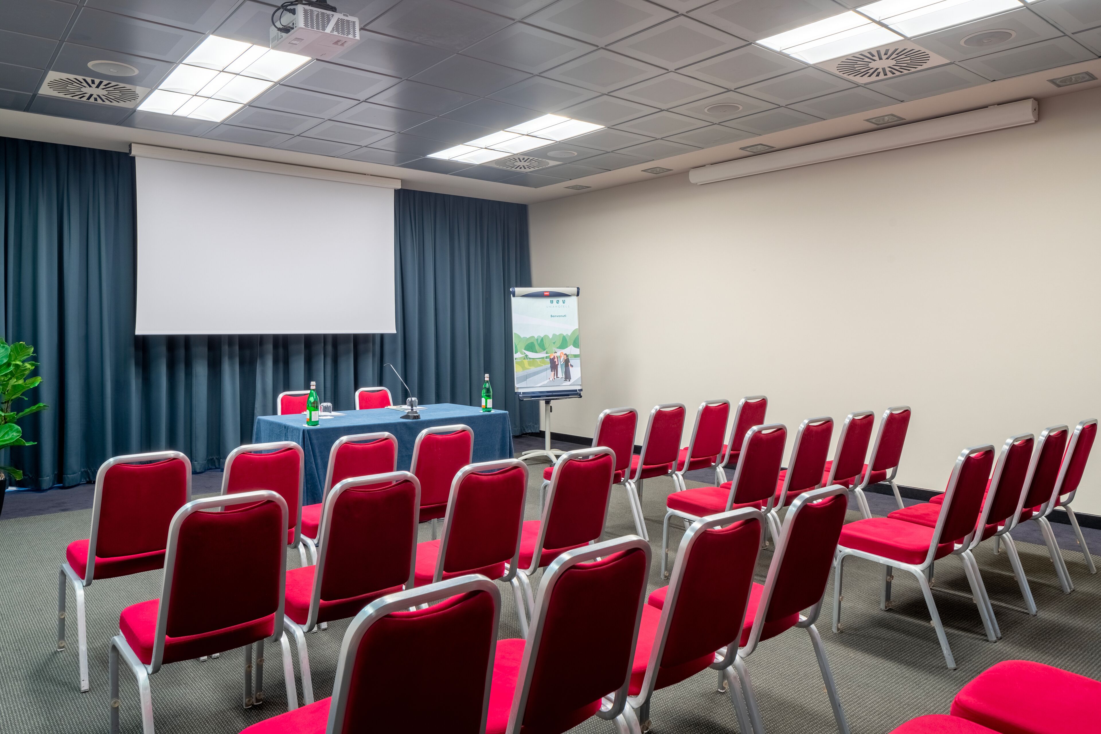 Una sala de conferencias con sillas rojas, una mesa con pantalla para proyector y un cartel.