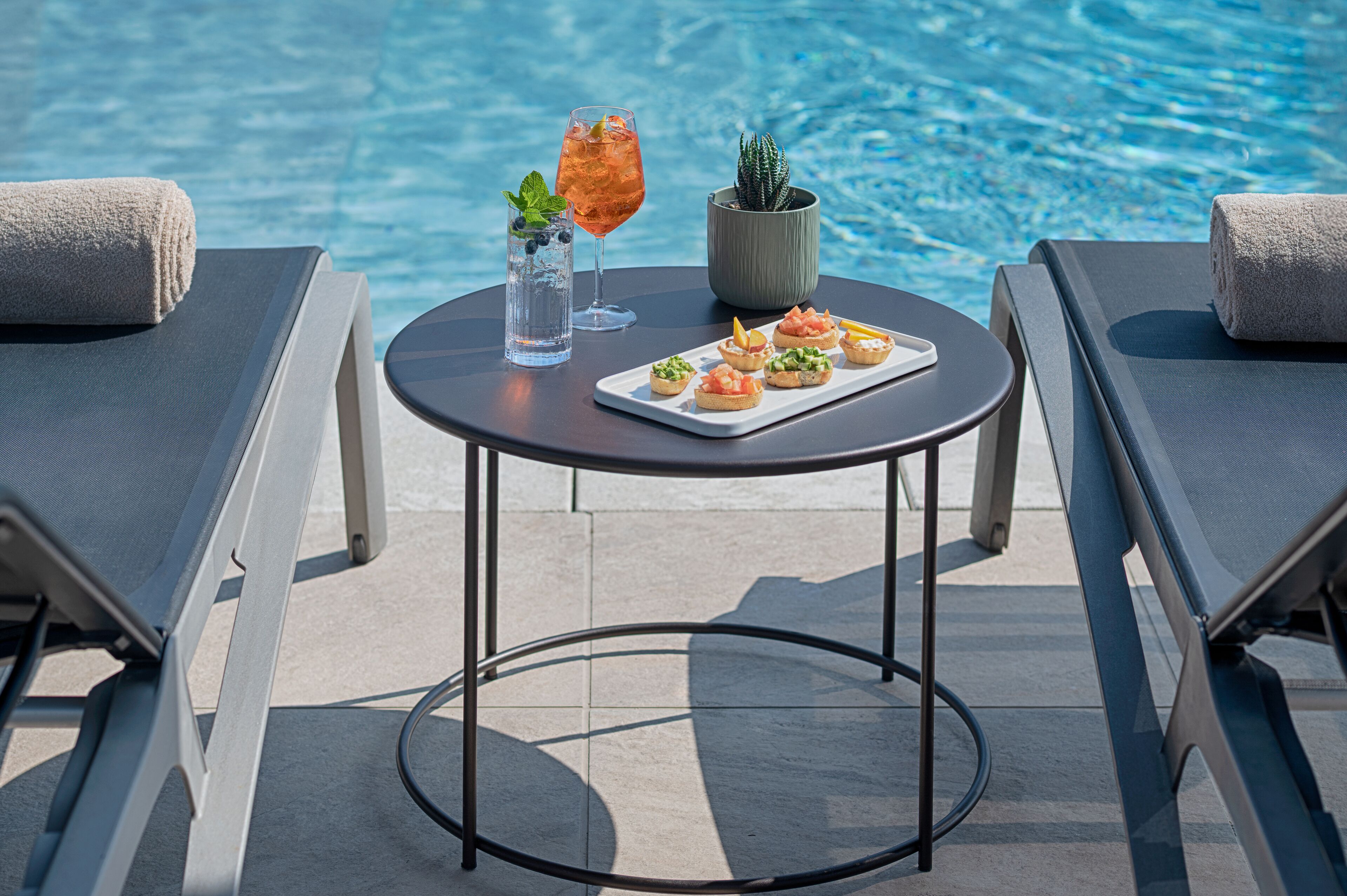 Mesa junto a la piscina con una bandeja de aperitivos, un vaso de bebida helada, una planta en maceta y toallas en sillones de descanso.