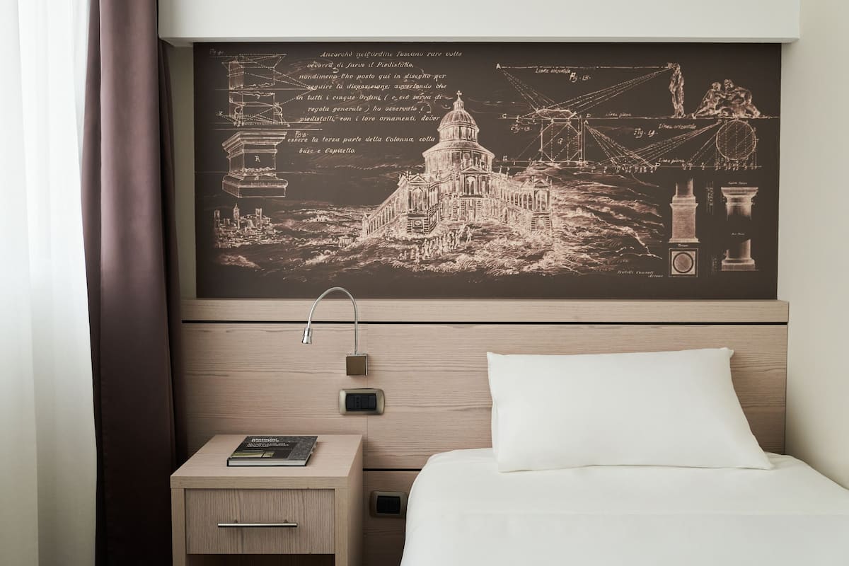 Ein Hotelzimmer mit einem Bett, einem Nachttisch mit einem Buch, einer Wanduhr und einem großen gerahmten Bild eines Gebäudes.