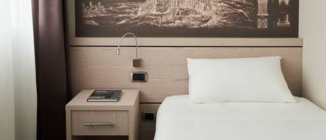 A hotel room with a bed, a nightstand with a book, a wall-mounted clock, and a large framed picture of a building.