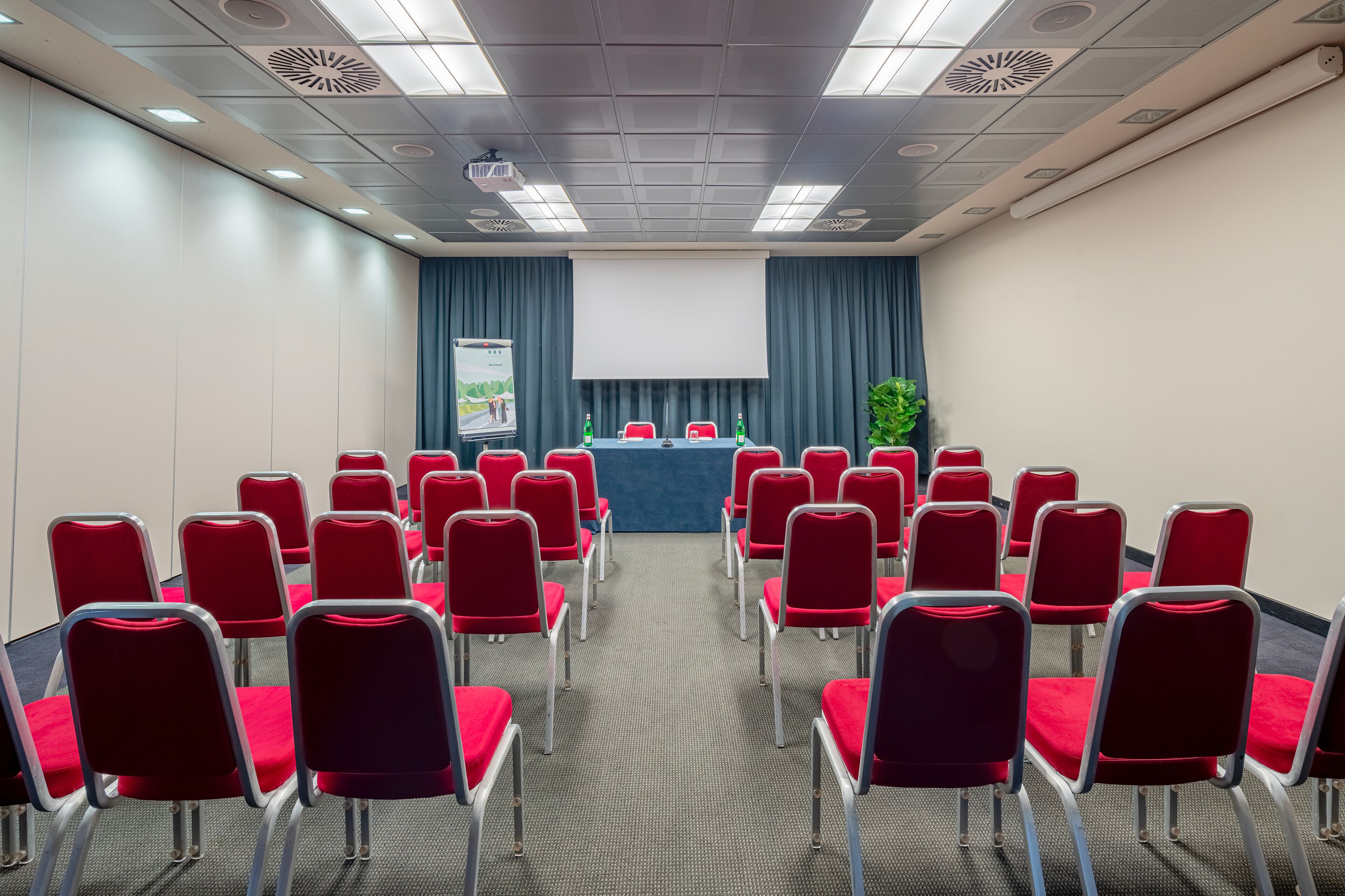 Un salón de conferencias con filas de sillas rojas mirando una pantalla de presentación, un atril y un proyector.