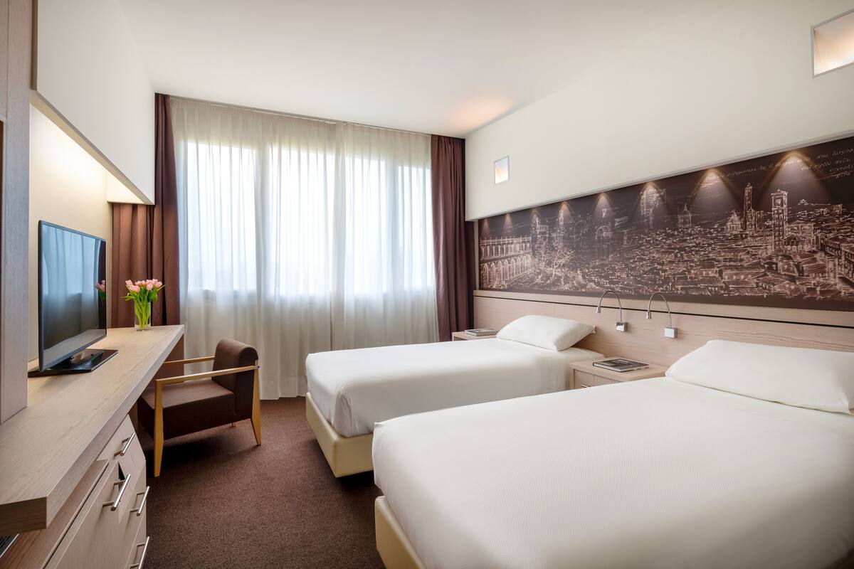 Ein Hotelzimmer mit zwei Betten, einem Schreibtisch mit Fernseher, einem Sessel und einem großen Wandgemälde mit Stadtansicht.