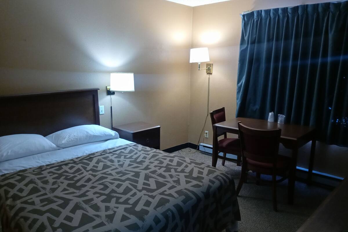 A hotel room with a bed, bedside tables, a desk with a chair, and a window with curtains.