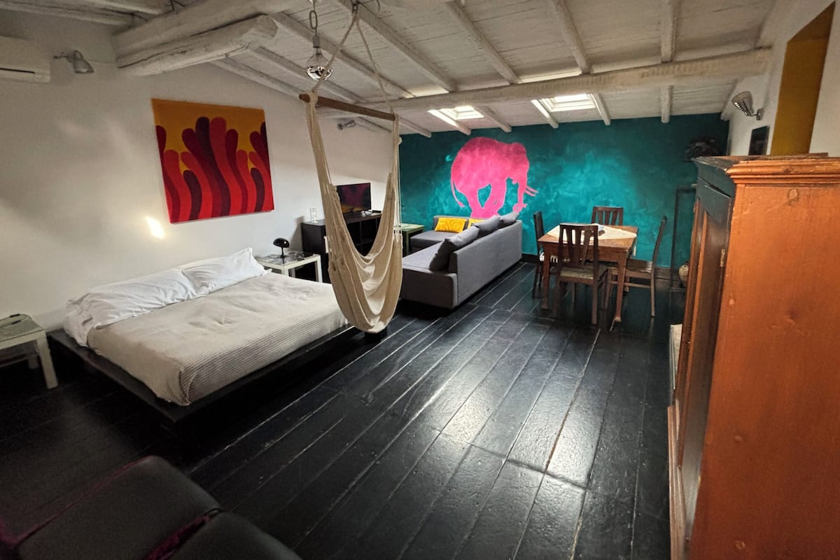 A room with a bed, a hanging chair, a sofa, a dining table, and a wall mural.