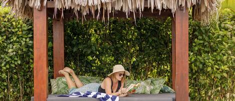 A poolside lounge with a thatched roof, a cushioned bench, and a person reading a book.