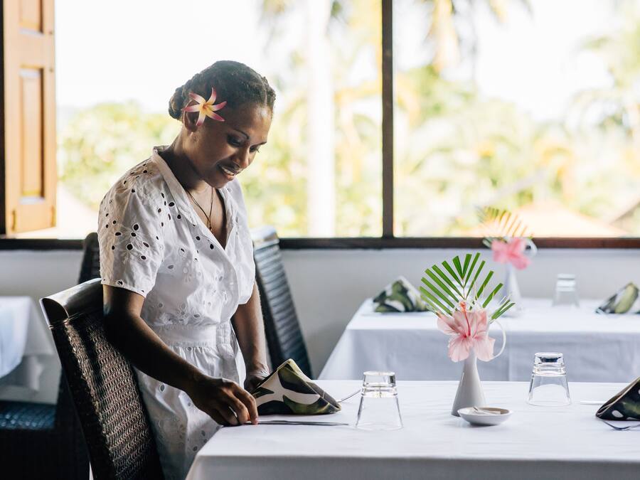 A person seated at a table with a white tablecloth, a vase with pink flowers, and a small glass container.
