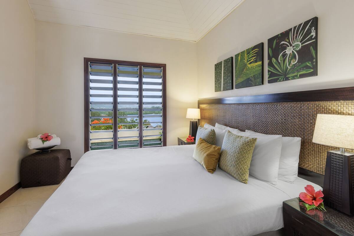 A hotel room with a large bed, a nightstand with a lamp, a small table with a flower, and a view of the outdoors through a sliding door.