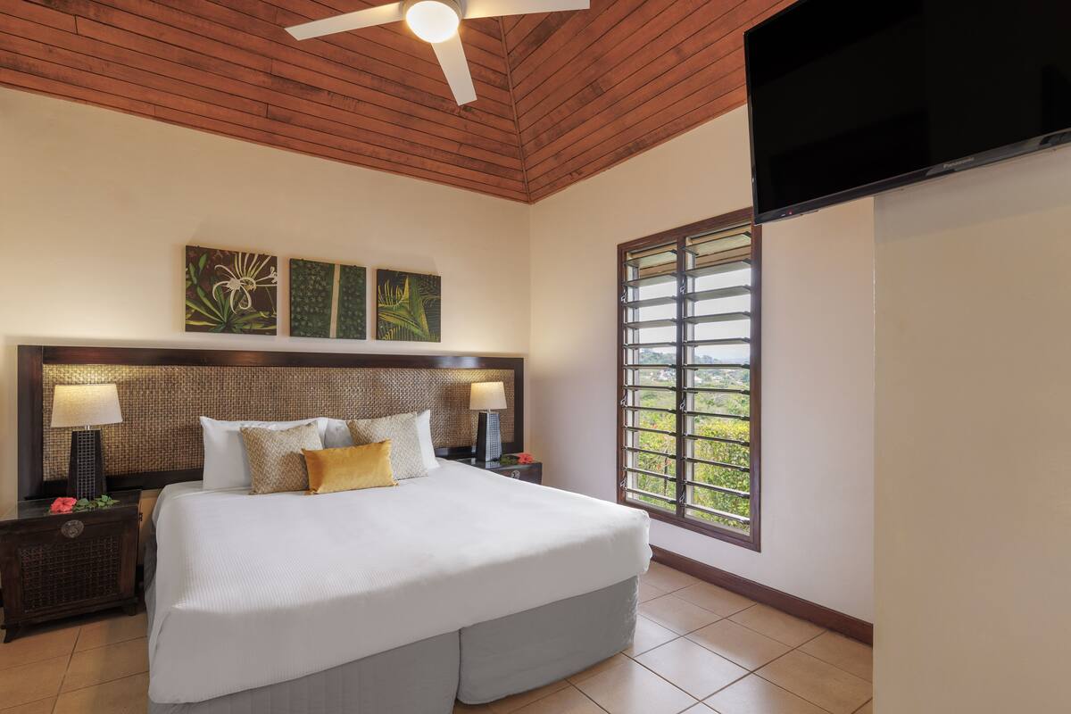 A spacious bedroom with a large bed, wooden ceiling, and a window with a view of greenery.