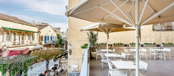 Un café al aire libre con mesas blancas y sombrillas, un edificio de piedra con contraventanas azules y una calle con gente caminando.