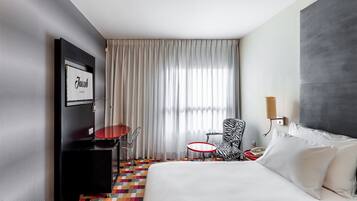 Habitación de hotel con cama, escritorio, silla, mesita, lámpara y ventana grande con cortinas.