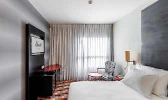 Habitación de hotel con cama, escritorio, silla, mesita, lámpara y ventana grande con cortinas.