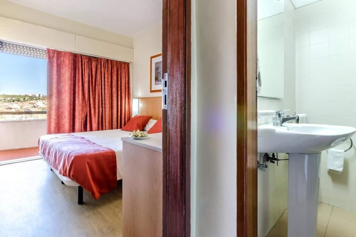 Habitación de hotel con cama, mesita de noche, ventana con cortinas, baño con lavamanos y vistas a la ciudad.