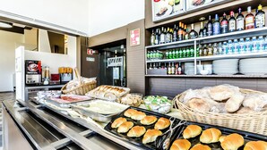Zona de bufé con diversas opciones de comida, como pan y bollos, y una selección de bebidas en estantes.