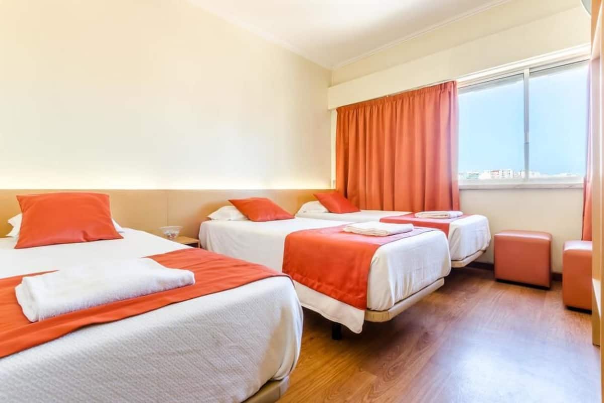 Habitación de hotel con tres camas, cada una con ropa blanca y detalles naranja, una ventana con cortinas naranja y suelo de madera.