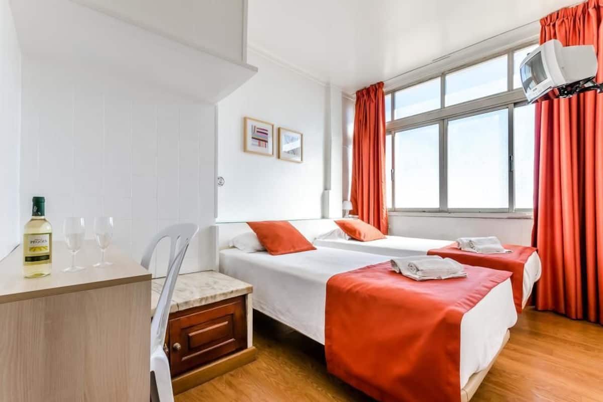 Habitación de hotel con dos camas, un escritorio con una botella y vasos, un televisor y una ventana con cortinas.