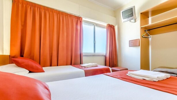 Habitación de hotel con dos camas, cortinas naranjas, un televisor montado en la pared y un estante de madera con una percha.