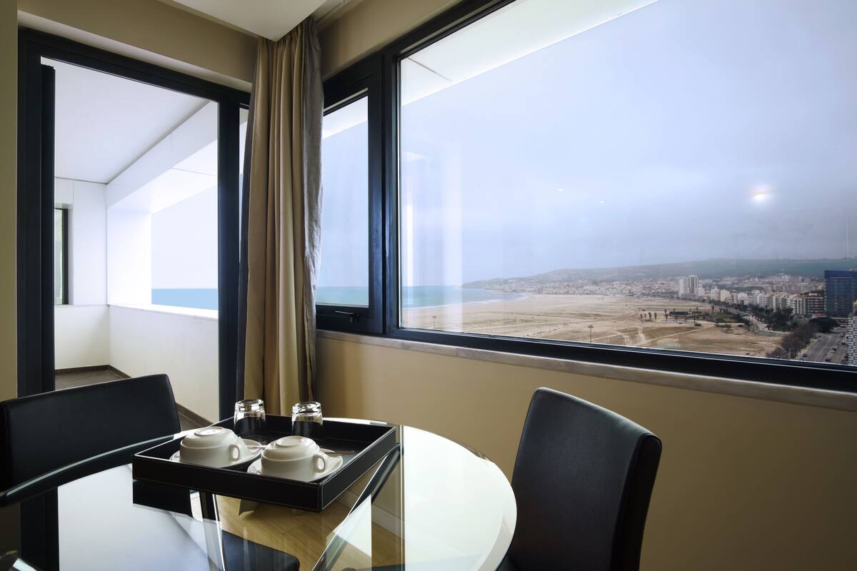 A hotel room with a glass table, two chairs, a tray with cups and a small container, and a view of a cityscape through large windows.