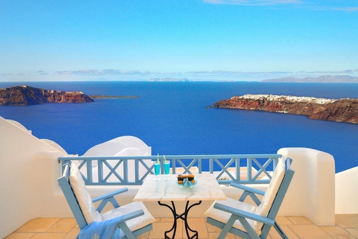 A terrace with a table and chairs overlooking a blue sea and distant land.