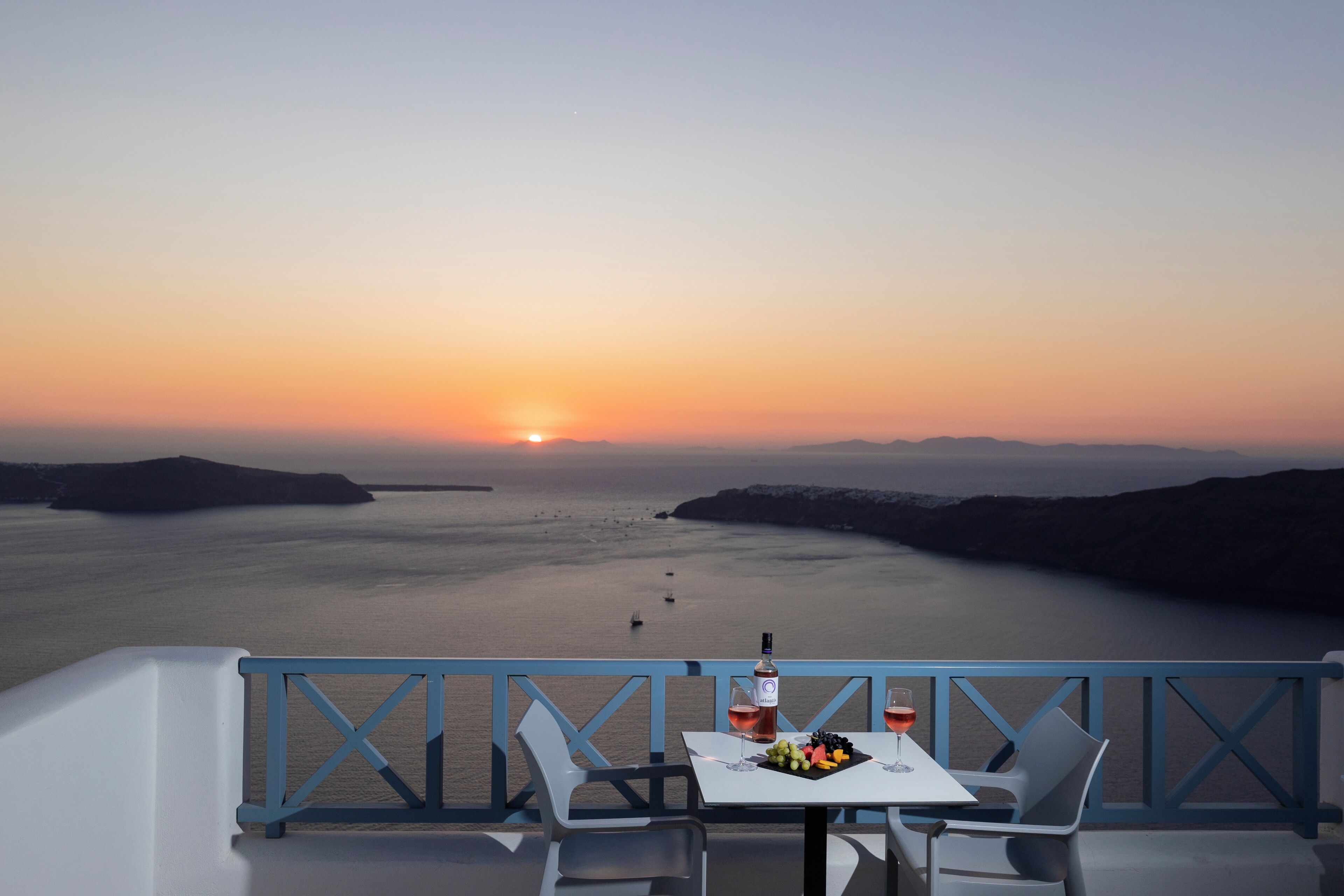 Ein Tisch mit einer Flasche und Gläsern, ein Teller mit Obst und Blick auf das Meer bei Sonnenuntergang.