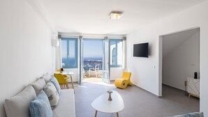 A modern living room with a sofa, a small table, and a yellow chair. There is a TV mounted on the wall and a door leading to an outdoor area with a view of the sea. - Absolute Bliss (Santorini)