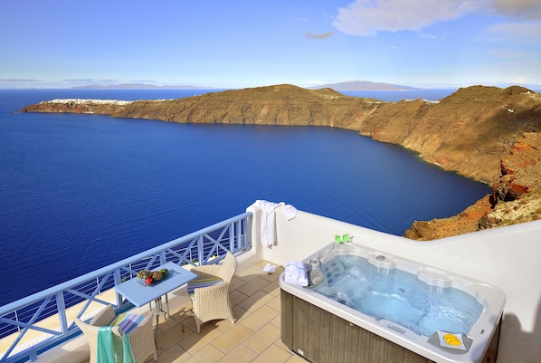 A balcony with a hot tub, a table with fruits, and a chair, overlooking a coastal landscape with cliffs and the sea. - Absolute Bliss (Santorini)