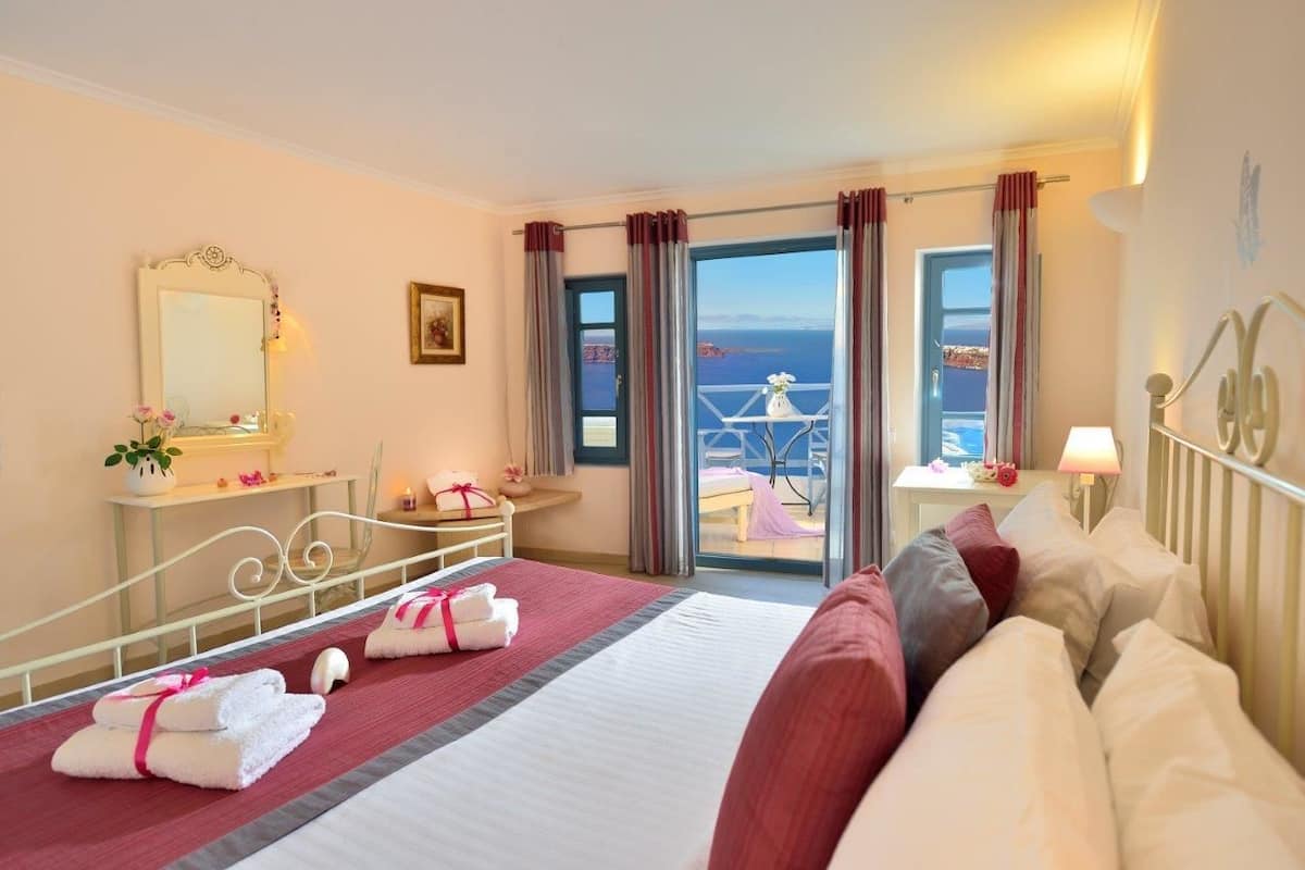 A hotel room with a bed, a desk with a lamp, a mirror, and a view of the sea through large windows.